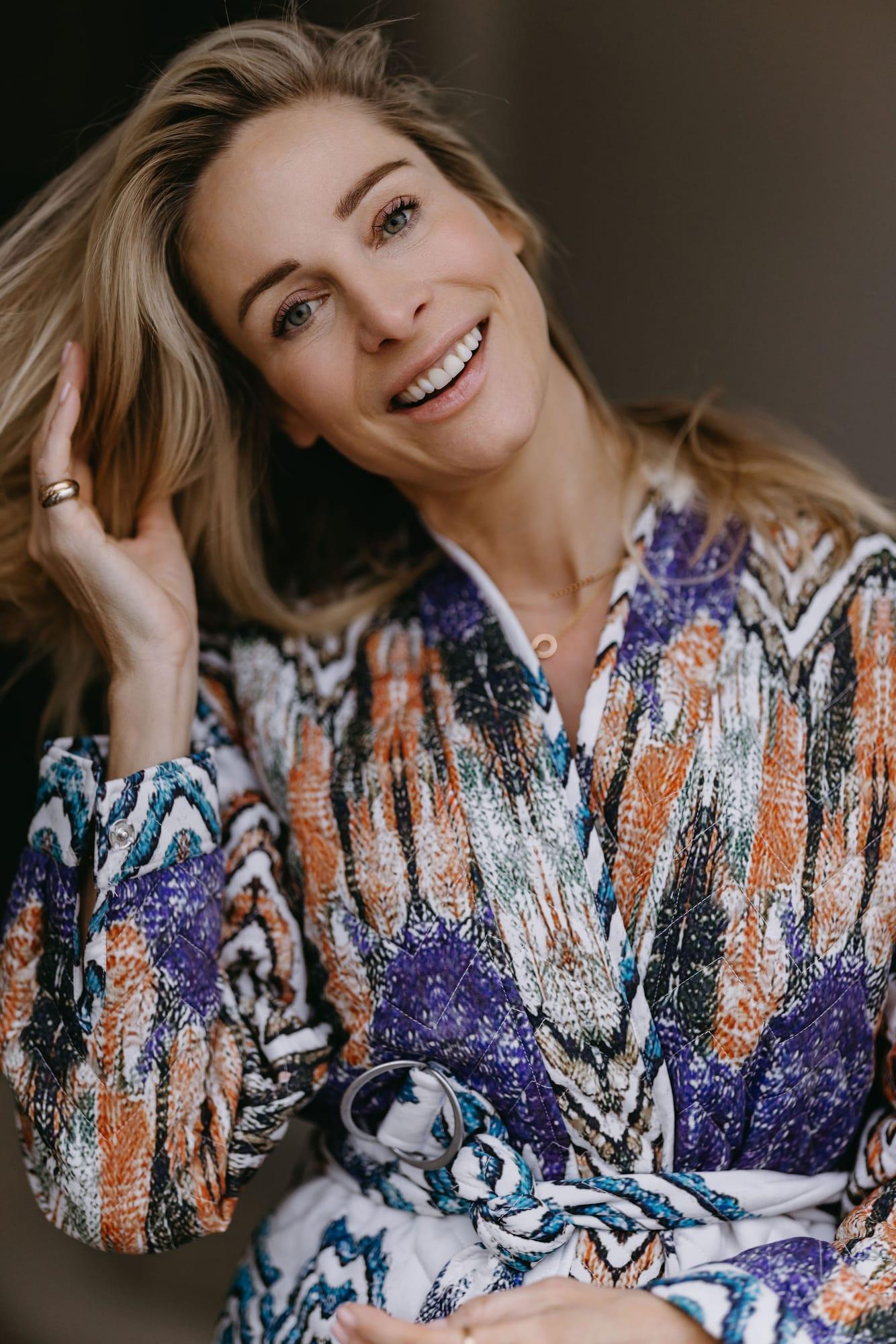 Virginie Versteegh in een kleurrijke blouse, lachend met een hand in haar haar