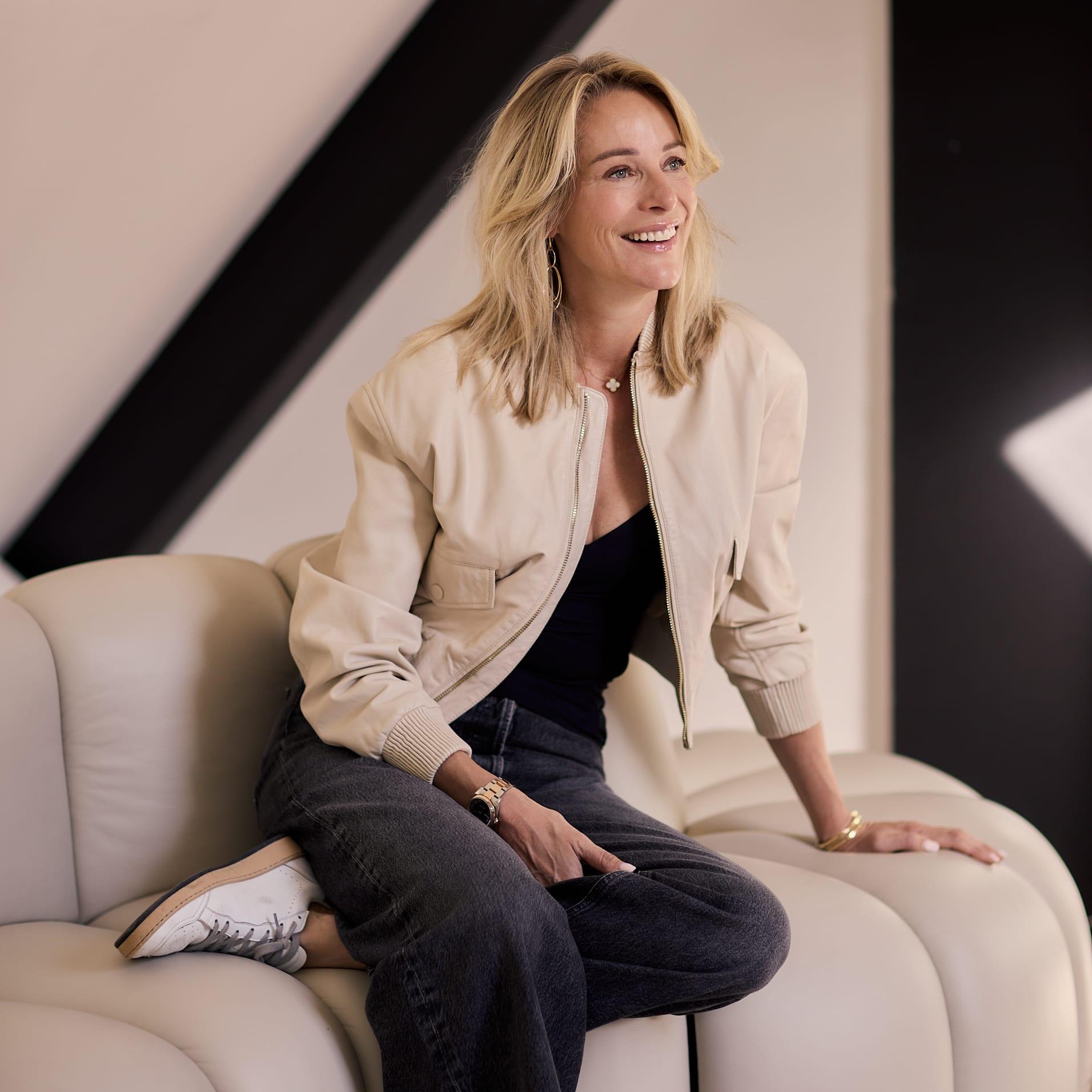 Virginie Versteegh in een beige leren jack, lachend zittend op een lichte bank