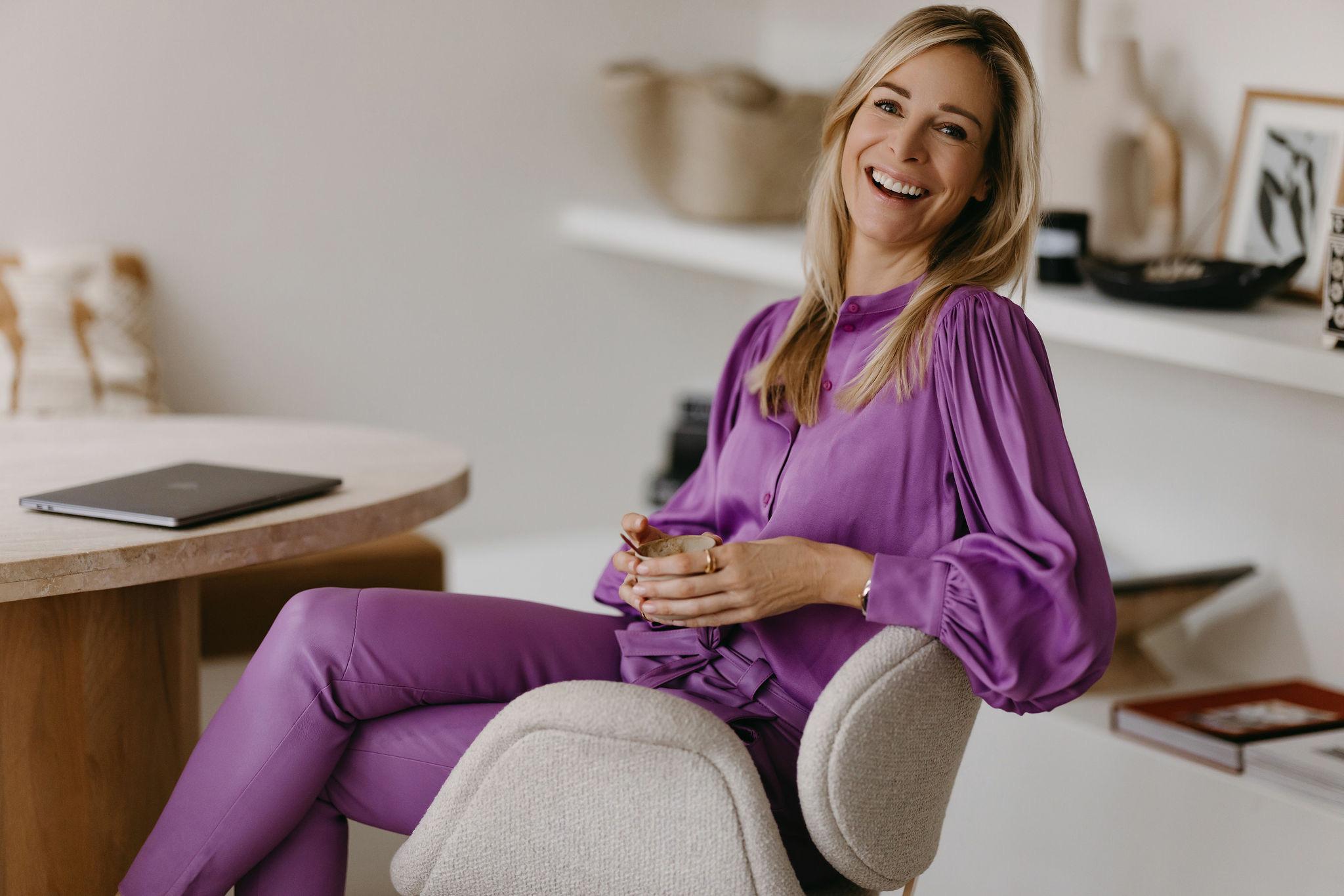 Virginie Versteegh in een paars pak, lachend op een stoel met een kop koffie